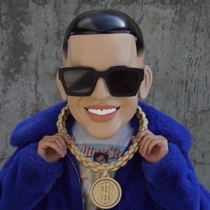 May include: A blue faux fur jacket with a gold chain necklace and a large gold dollar sign pendant. The person wearing the jacket is wearing a black mask with sunglasses and a white t-shirt with the text "Billionaire Status" printed on it.