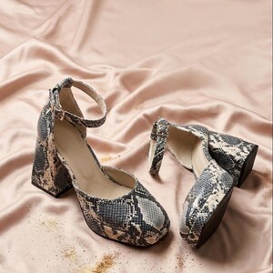 May include: A pair of snakeskin-patterned block heel pumps with ankle straps. The shoes feature a rounded toe and a chunky heel. The background is a satin fabric in a light pink color.