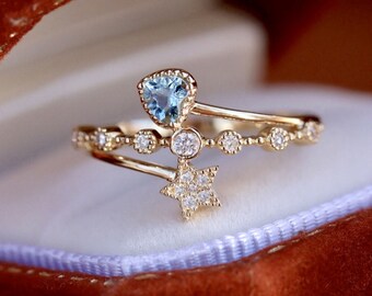 May include: A gold-colored ring featuring a light blue heart-shaped gemstone and a star-shaped cluster of smaller stones. The ring has a delicate design with small, round, clear stones along the band, set against a white background.
