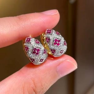 May include: A pair of gold hoop earrings with a floral design. The earrings are set with small red and white gemstones.