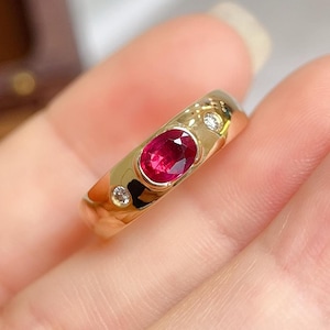 May include: A gold ring with an oval ruby gemstone and two small diamonds. The ring's polished surface reflects light. The ruby is the focal point, set in a gold band. The ring is presented against a soft background.