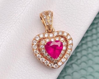 Puede incluir: Un colgante en forma de corazón con una vibrante gema roja en el centro, rodeado por un doble halo de pequeñas piedras blancas brillantes. El colgante está engastado en metal dorado y tiene una fianza decorativa.