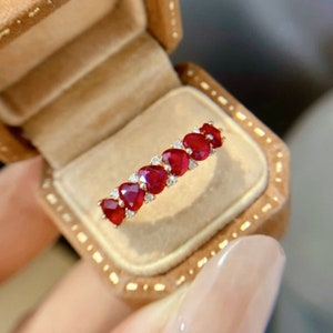 Pode incluir: Um anel de banda de ouro com uma fileira de pedras de rubi em forma de coração engastadas na banda. O anel está em uma caixa de anel de veludo.