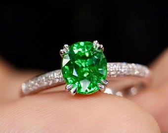 May include: A silver ring with a large, oval-cut, bright green gemstone. The band is set with small, sparkling stones. The ring is shown against a dark background, emphasising the gemstone's colour and sparkle. The ring is a piece of jewellery.