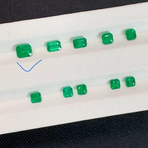 May include: A selection of emerald-cut emerald gemstones, each with a vivid green colour. The gemstones are displayed on a white surface, highlighting their clarity and facets. The emeralds vary slightly in size and shape.