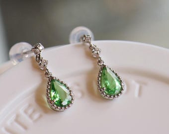 May include: A pair of silver drop earrings featuring pear-shaped, vibrant green gemstones. The earrings have a delicate design with small, clear accent stones and a beaded border around the green gems. The earrings are displayed on a white surface.