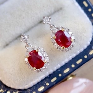 Puede incluir: Un par de pendientes colgantes con piedras preciosas de rubí de forma ovalada rodeadas por un halo de diamantes más pequeños. Los pendientes son de plata y tienen un acabado pulido.