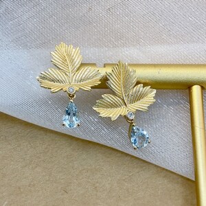 May include: Gold leaf earrings with a teardrop shaped aquamarine gemstone dangling from each leaf. The leaves are textured and have a delicate design.