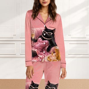May include: Pink pajama set featuring a black cat wearing sunglasses and a pearl necklace, lounging on a pink couch with a glass of champagne. The set includes a button-down top with black trim and matching pants.