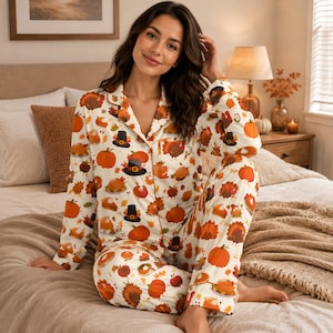 Op de afbeelding: Een tweedelige pyjamaset bestaande uit een shirt met lange mouwen en knoopsluiting en een bijpassende broek. De witte stof is voorzien van een herhalend patroon van oranje pompoenen, bruine kalkoenen, zwarte hoeden en stukjes pompoentaart.