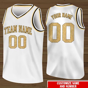 Puede incluir: Camiseta de baloncesto blanca con ribetes negros y dorados. La parte delantera de la camiseta tiene el texto "TEAM NAME" y el n&uacute;mero "00" en purpurina dorada. La parte trasera de la camiseta tiene el texto "YOUR NAME" y el n&uacute;mero "00" en purpurina dorada.
