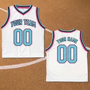 May include: White basketball jersey with black and pink trim. The front of the jersey has the text "YOUR TEAM" and the number "00" in light blue. The back of the jersey has the text "YOUR NAME" and the number "00" in light blue.