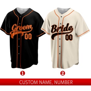 May include: Two baseball jerseys, one black with "Groom 00" in orange text and one cream with "Bride 00" in orange text. Both jerseys have orange piping and buttons.