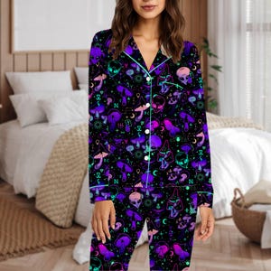 Peut inclure: Ensemble de pyjama noir avec un motif vibrant de champignons violets, de t&ecirc;tes d'extraterrestres et de symboles c&eacute;lestes. Le haut &agrave; manches longues et le pantalon ont une bordure turquoise et des boutons blancs. Le pyjama est en mati&egrave;re douce et confortable.
