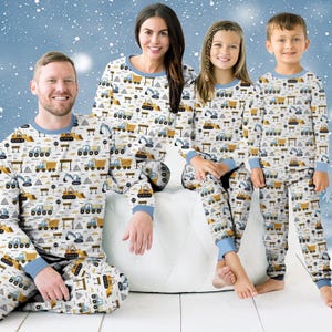 May include: Family pyjamas with a construction vehicle pattern in yellow, blue, and grey on a white background. The tops and trousers have blue trim. The family is sitting on a white cushion. The pyjamas are for adults and children.