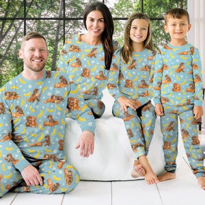 Puede incluir: Pijamas familiares a juego en azul claro con un patr&oacute;n repetido de monos marrones, pl&aacute;tanos amarillos y estrellas blancas. Los pijamas incluyen tops y pantalones de manga larga. La familia est&aacute; sentada en una superficie blanca.