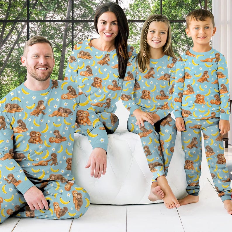 Puede incluir: Pijamas familiares a juego en azul claro con un patr&oacute;n repetido de monos marrones, pl&aacute;tanos amarillos y estrellas blancas. Los pijamas incluyen tops y pantalones de manga larga. La familia est&aacute; sentada en una superficie blanca.