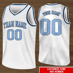 Puede incluir: Camiseta de baloncesto blanca con ribetes azules y negros. La parte delantera de la camiseta tiene el texto "TEAM NAME" y el n&uacute;mero "00" en azul con un efecto brillante. La parte trasera de la camiseta tiene el texto "YOUR NAME" y el n&uacute;mero "00" en azul con un efecto brillante.