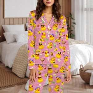 May include: Pink pajama set with a repeating pattern of yellow ducks wearing sunglasses, hats, and holding tropical drinks. The pajamas have white trim and a button-down top. The background has a textured appearance.