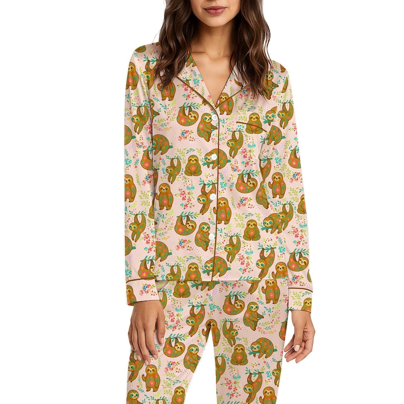 May include: A light pink pajama set featuring a repeating pattern of sloths and floral elements. The long-sleeved top and pants have brown trim. The sloths are in various poses, adding a playful touch to the sleepwear.