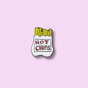 May include: A white enamel pin depicting a bag of hot chips. The bag has the words "HOT CHIPS" in red lettering. The chips are yellow and protrude from the top. The pin has a black outline and is set against a pink background.