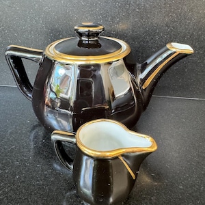 Könnte beinhalten: Schwarzes Keramik-Teeservice mit goldenen Verzierungen. Die Teekanne hat einen Deckel und einen gebogenen Ausguss. Der Sahnekännchen hat einen Griff und einen Ausguss. Beide Teile haben ein glänzendes Finish.