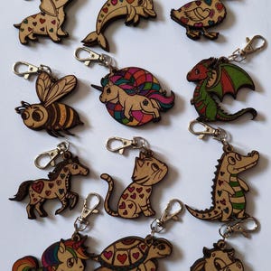 Puede incluir: Un conjunto de 12 llaveros de madera con diferentes diseños de animales. Cada llavero está pintado con detalles coloridos y tiene un anillo de metal plateado. Los animales incluyen un unicornio, un gato, un dragón, una abeja, una tortuga, un caballo, un delfín, un pato, un cocodrilo, un dachshund y una vaca.