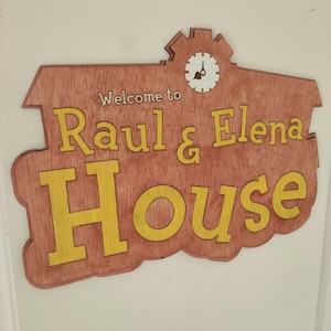 Puede incluir: Un letrero de madera con un esquema de color amarillo y marrón. El letrero dice "Bienvenido a Raul & Elena House" con una esfera de reloj en la esquina superior izquierda.