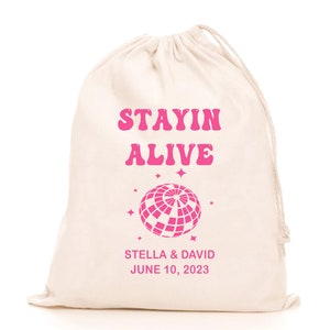 May include: White cotton drawstring bag with pink text that reads "Stayin Alive" with a pink disco ball graphic and the text "Stella & David June 10, 2023".