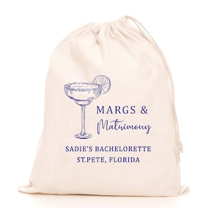 May include: A cream-coloured drawstring bag featuring a blue margarita glass illustration and text. The text reads "MARGS & Matrimony" and "SADIE'S BACHELORETTE ST. PETE, FLORIDA". The bag has a drawstring closure.