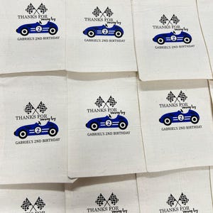K&ouml;nnte beinhalten: Eine Sammlung von beigefarbenen Baumwollbeuteln mit Kordelzug, bedruckt mit einer blauen Rennwagen-Grafik mit der Nummer zwei, zwei Zielflaggen und dem Text: THANKS FOR racing by GABRIEL'S 2ND BIRTHDAY. Die Beutel sind in einem Raster angeordnet.