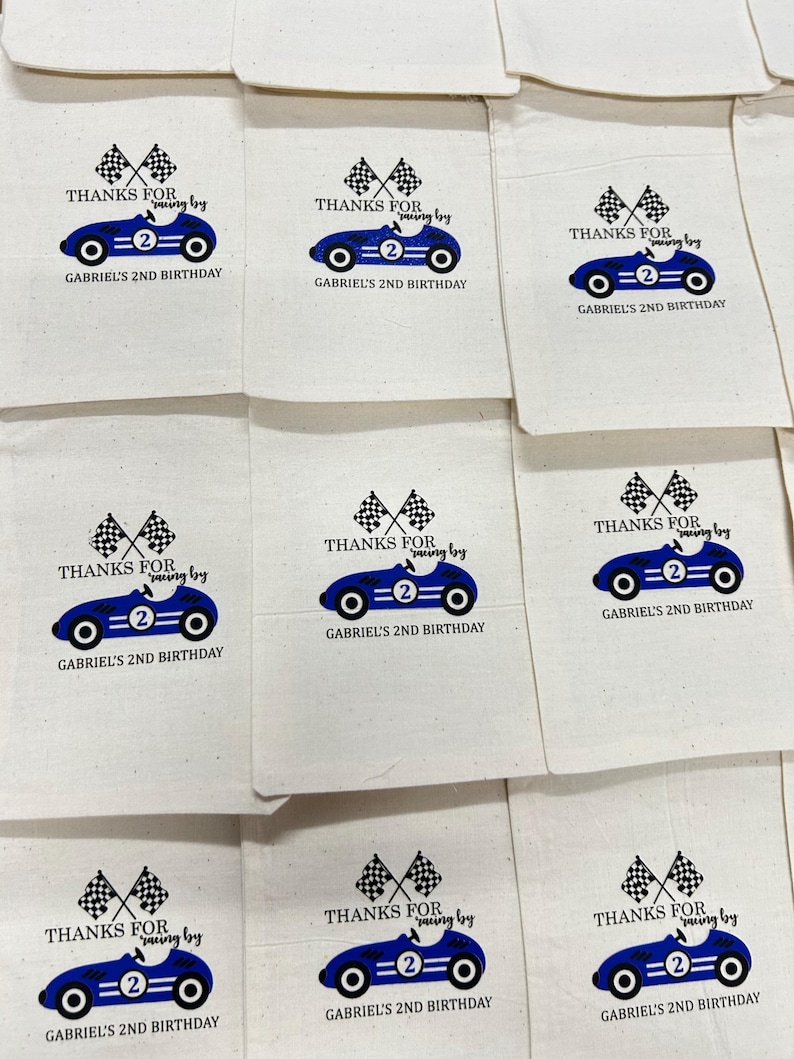 K&ouml;nnte beinhalten: Eine Sammlung von beigefarbenen Baumwollbeuteln mit Kordelzug, bedruckt mit einer blauen Rennwagen-Grafik mit der Nummer zwei, zwei Zielflaggen und dem Text: THANKS FOR racing by GABRIEL'S 2ND BIRTHDAY. Die Beutel sind in einem Raster angeordnet.