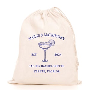 May include: A white cotton drawstring bag with blue text and a blue illustration of a margarita glass. The text reads "Margs & Matrimony Est. 2024 Sadie's Bachelorette St. Pete, Florida."
