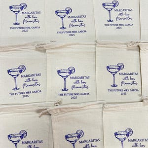 K&ouml;nnte beinhalten: Eine Sammlung von cremefarbenen Stoffbeuteln mit Kordelzug, bedruckt mit einer blauen Grafik eines Margaritaglases mit Limettenscheibe. Der Text lautet: MARGARITAS with her Mamasitas THE FUTURE MRS. GARCIA 2025. Die Beutel sind in einem Raster angeordnet.