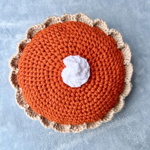 May include: A crocheted pumpkin pie with a white dollop of whipped cream. The pie has an orange filling and a tan crust with a scalloped edge. The handmade pie is a decorative item.