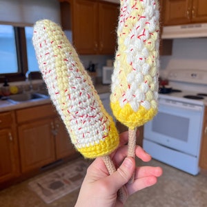 Puede incluir: Dos juguetes de maíz en la mazorca hechos a crochet. El maíz es amarillo y blanco con costuras rojas. El maíz está unido a un palo marrón.