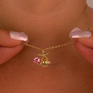 Peut inclure: Un d&eacute;licat collier en or avec un pendentif orn&eacute; d'une pierre rose et d'un motif coquillage. Le collier est tenu par deux mains. Le pendentif mesure environ 1,3 cm de long.