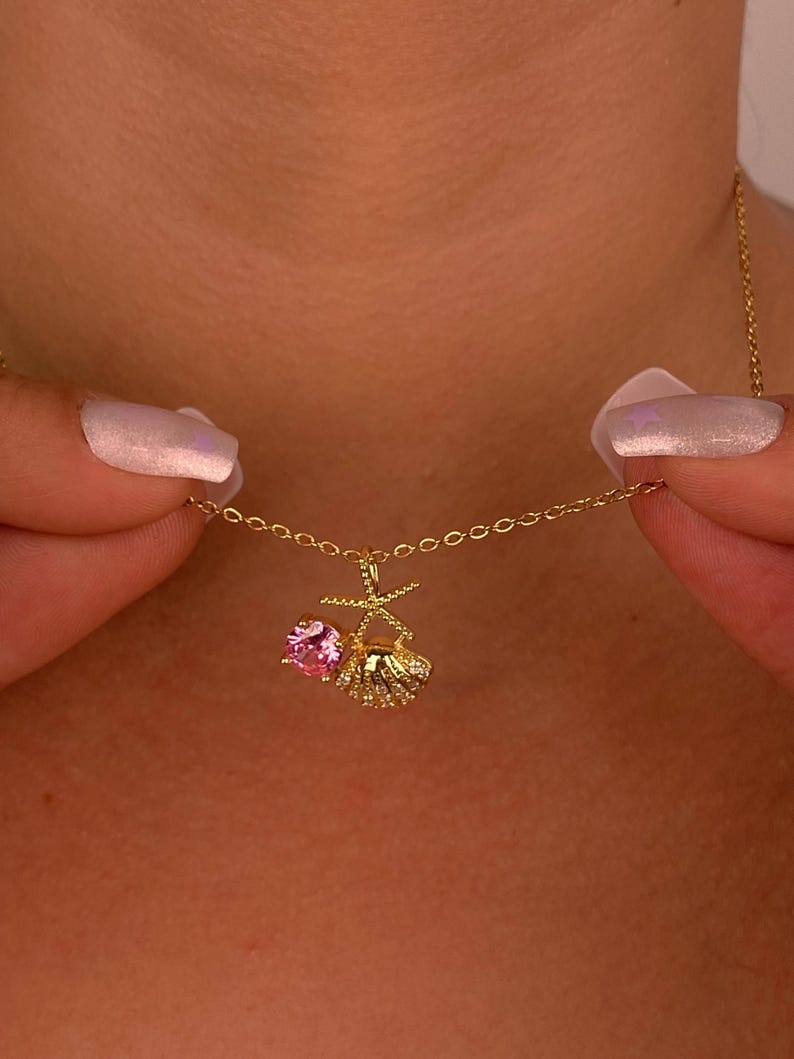 Peut inclure: Un d&eacute;licat collier en or avec un pendentif orn&eacute; d'une pierre rose et d'un motif coquillage. Le collier est tenu par deux mains. Le pendentif mesure environ 1,3 cm de long.