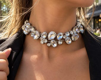 May include: A choker necklace featuring multiple clear, faceted crystals in various shapes and sizes, including pear and oval cuts, set in a silver-toned metal frame that sits closely around the base of the neck.