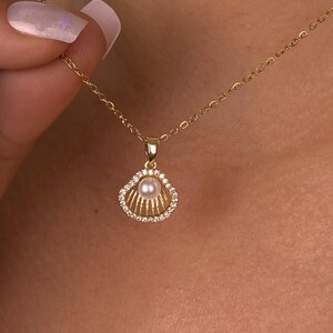Op de afbeelding: Een goudkleurige ketting met een schelpenhanger. De hanger heeft een parel in het midden, omringd door kleine, heldere edelstenen. De ketting is delicaat en ook goudkleurig. De ketting wordt door een persoon vastgehouden.