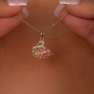 Peut inclure: Un collier avec un pendentif en forme de cygne dor&eacute;. Le cygne est orn&eacute; de pierres pr&eacute;cieuses claires et roses, et le collier est tenu par une personne. Le corps du cygne est fait de pierres roses, et les ailes sont faites de pierres claires et roses.