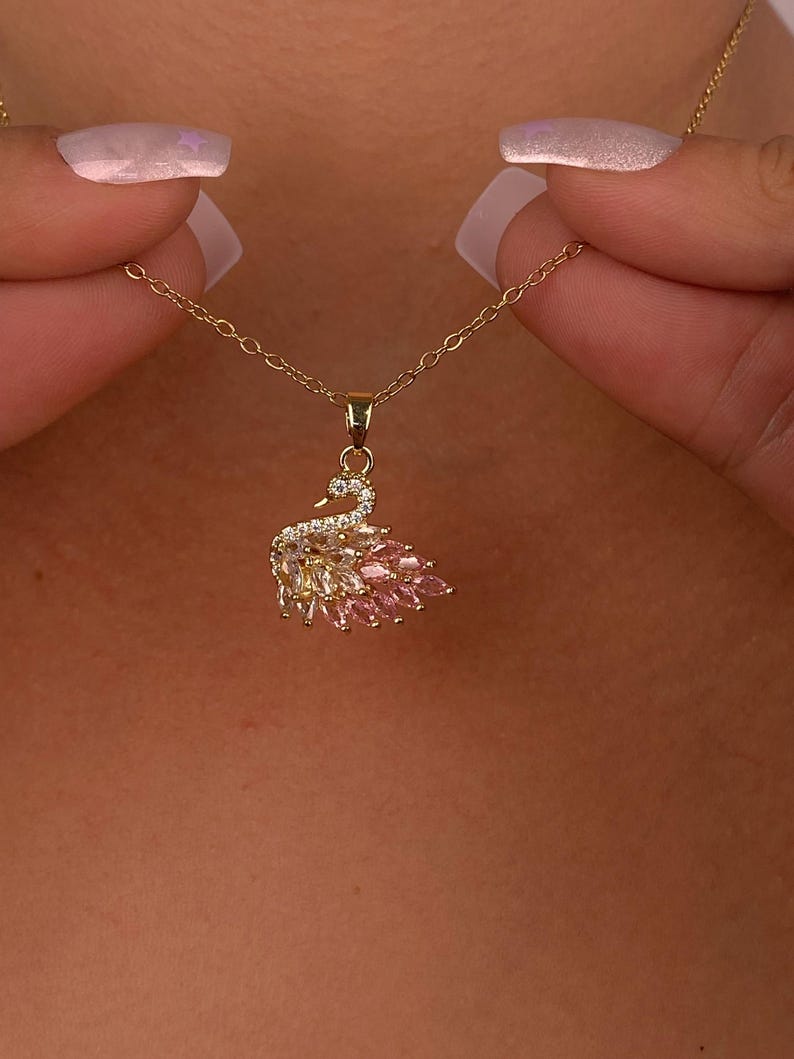 Peut inclure: Un collier avec un pendentif en forme de cygne dor&eacute;. Le cygne est orn&eacute; de pierres pr&eacute;cieuses claires et roses, et le collier est tenu par une personne. Le corps du cygne est fait de pierres roses, et les ailes sont faites de pierres claires et roses.