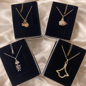 Peut inclure: Quatre colliers dor&eacute;s avec diff&eacute;rents pendentifs : une cerise, un cygne, un squelette de poisson et une forme d'&eacute;ventail. Chaque collier est pr&eacute;sent&eacute; dans un coffret noir sur un fond satin&eacute;.