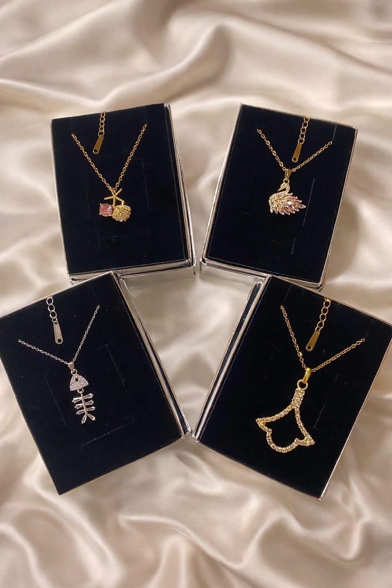 Peut inclure: Quatre colliers dor&eacute;s avec diff&eacute;rents pendentifs : une cerise, un cygne, un squelette de poisson et une forme d'&eacute;ventail. Chaque collier est pr&eacute;sent&eacute; dans un coffret noir sur un fond satin&eacute;.