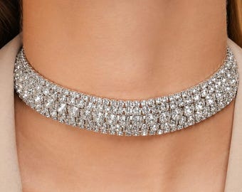 May include: A multi-row choker necklace featuring three horizontal bands of clear, round-cut rhinestones set in a silver-toned metal frame. The design consists of small, tightly packed crystals that create a continuous, reflective, and sparkling surface.