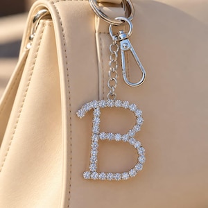 May include: A silver-toned metal keychain charm in the shape of the letter B, encrusted with clear, round-cut rhinestones. The charm is attached to a silver-colored metal chain and a lobster claw clasp, hanging from the handle of a beige handbag.