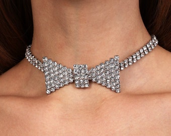 May include: A silver choker necklace featuring a large, sparkling rhinestone bow. The bow is made up of three sections, two triangular and one square.