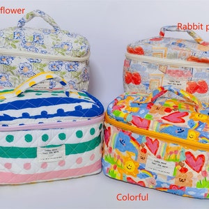 May include: Four quilted cosmetic bags with different patterns. One has blue floral print, another has a rabbit and postcard design, a third has blue stripes and green polka dots, and the last one is colorful with hearts and flowers. Each bag has a handle and a zipper.