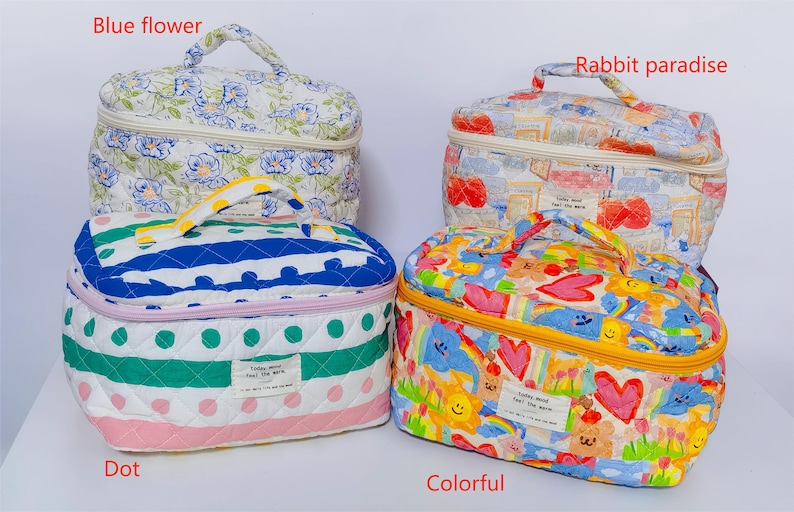 May include: Four quilted cosmetic bags with different patterns. One has blue floral print, another has a rabbit and postcard design, a third has blue stripes and green polka dots, and the last one is colorful with hearts and flowers. Each bag has a handle and a zipper.