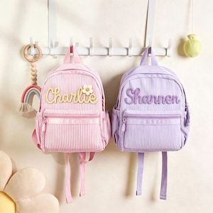 Könnte beinhalten: Zwei kleine Rucksäcke mit vertikalen Streifen, einer in Hellrosa und einer in Helllila. Der rosa Rucksack trägt den Namen Charlie in gelber Schrift mit einem Gänseblümchen, der lila Rucksack den Namen Shannen in lila Schrift.